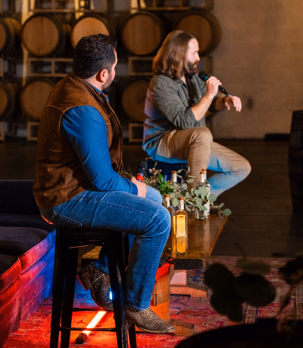 Two men on stools talking on a stage in front of barrels