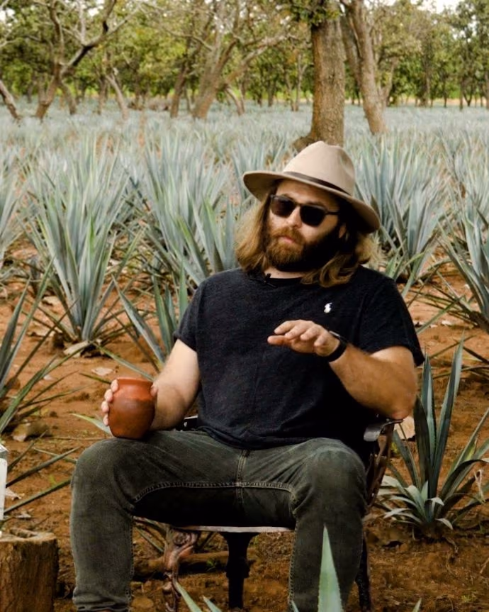Peter Kiley in a hat and sunglasses, seated among agave rows and holding a clay cup