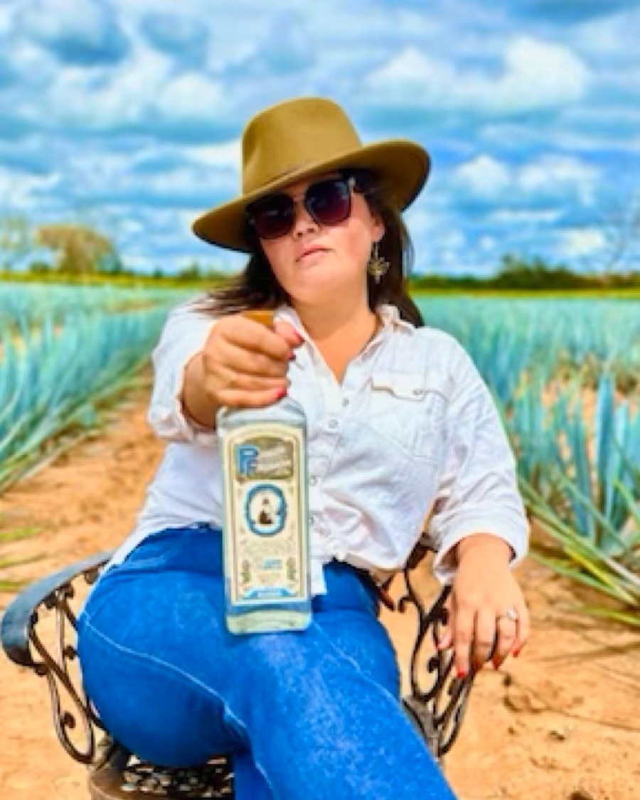 Rachel Kiley in a wide-brimmed hat and sunglasses, seated among blue agave and holding a bottle of Pedro Furtivo