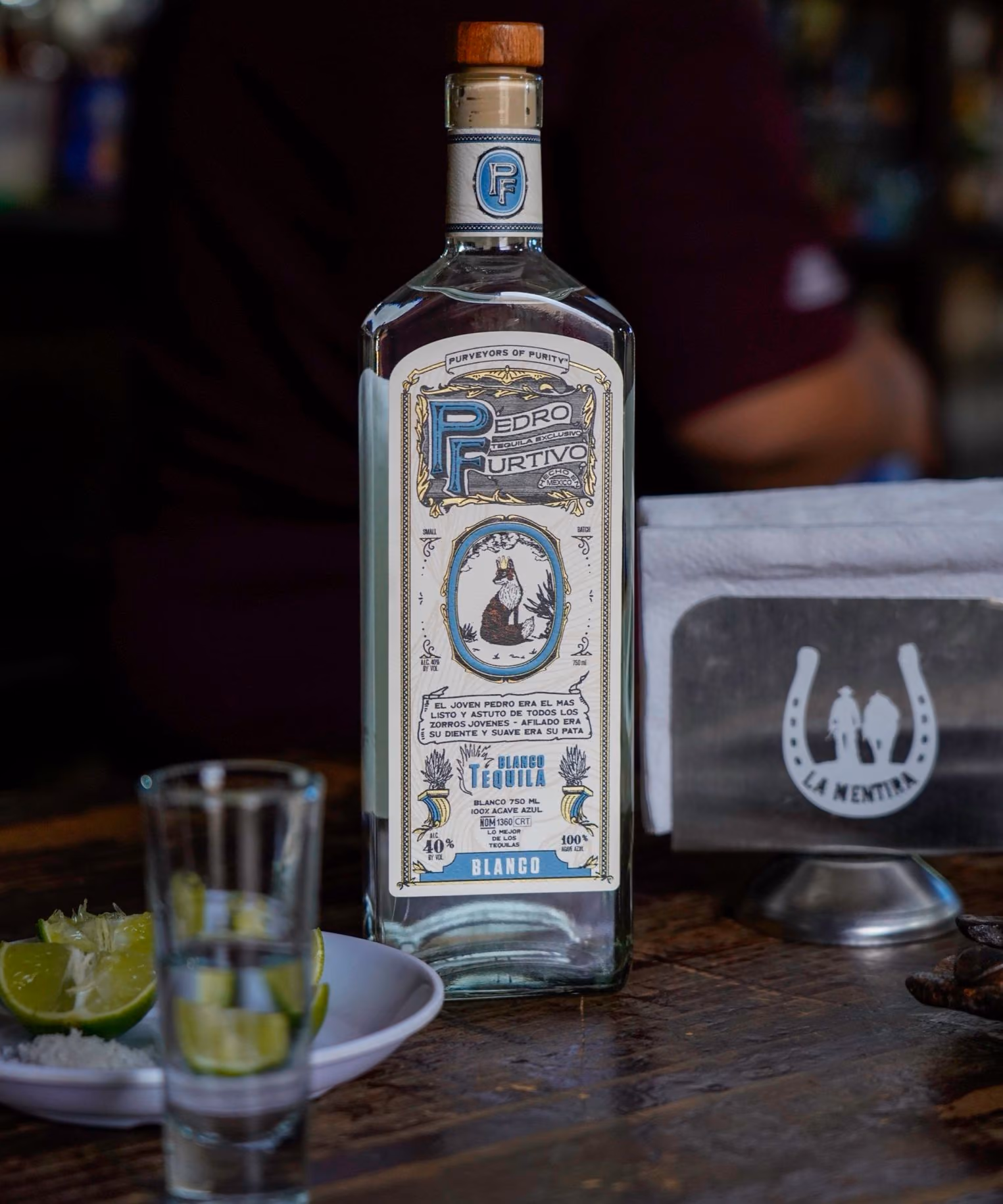 A second photograph of Pedro Furtivo Blanco, not its back label: the bottle on a dark bar table with a shot glass and limes