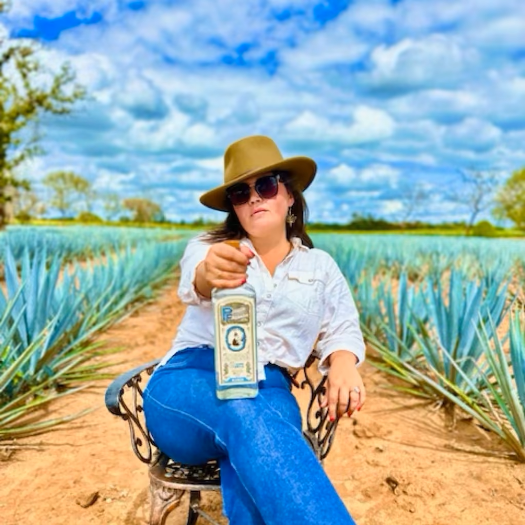 A woman in a straw hat seated in a field of blue agave, holding a bottle